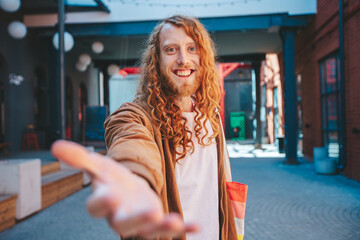 A happy smiling man with long curly red hair holding his hand out to the camera