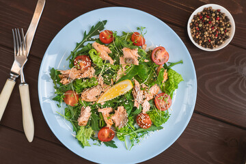 Salmon salad with green leaves, tomato, lemon, sesame in blue plate on wooden table