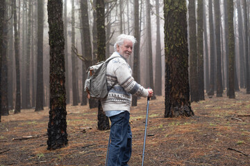 Active happy senior retired man with backpack walking in mountain forest on a foggy day with the help of poles enjoying nature, freedom and free time. Forest background with bare trees