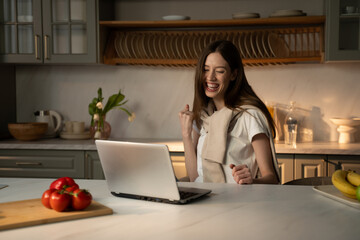 A young woman is seated at a kitchen counter with a laptop, her face lit up with a delighted smile as she appears to react to good news. The casual setting is highlighted by home details such as fresh