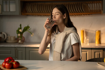 A young woman stands by a kitchen island, sipping water from a clear glass. Natural light pours in,...