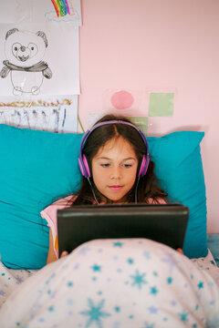 A little girl is looking at an electronic tablet lying in her bed 