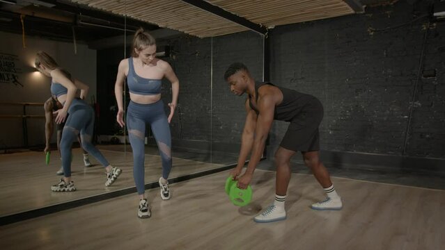 Motivated sporty muscular build black man practicing weight training with pretty female fitness trainer, performing bent over row with weight plates exercise at gym.