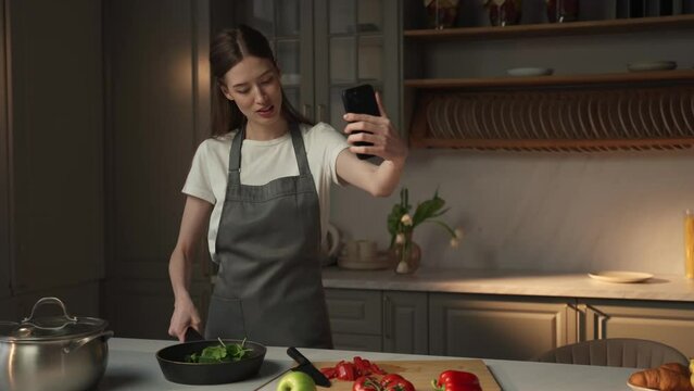 A young woman is standing in a contemporary kitchen wearing an apron, holding a frying pan in one hand and using a smartphone to record herself. She appears to be capturing a video tutorial, possibly