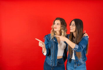 Point index finger aside, portrait of young female friends smiling point index finger aside. Showing copy space. Recommending concept idea. Standing together, wear denim jacket and jeans. 