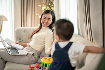 A happy Asian businesswoman mom is playing with her baby boy on a sofa while working from home.