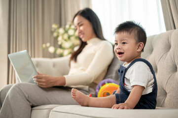 A cute Asian baby boy is playing with toys on a sofa while his mom is working on her laptop.