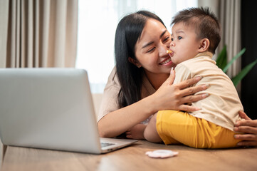 A happy Asian mom is playing and feeding her little son with snacks while working in her home office