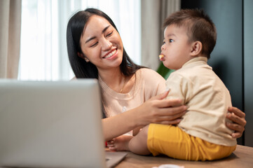 A happy Asian mom is playing and feeding her little son with snacks while working in her home office