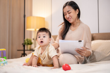 A happy, cute little son is playing with toys while his mom is working and reading documents in bed.