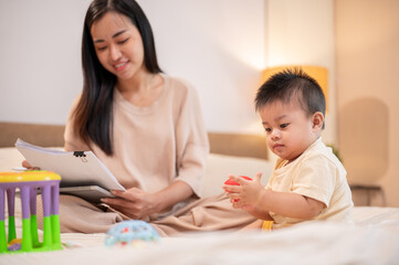 A happy, cute Asian baby boy is playing with toys while his mom is working in bed.