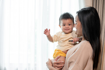 A caring Asian mom is holding her cute baby boy in her arms near by the window in the living room.