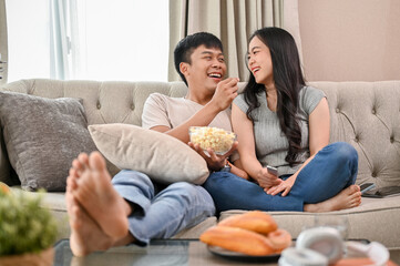 Obraz premium Happy young Asian couple enjoy eating popcorn and watching a movie while relaxing on a sofa.