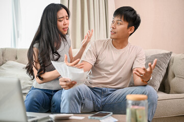 A resentful, angry Asian wife and husband are arguing about family household invoices on sofa.