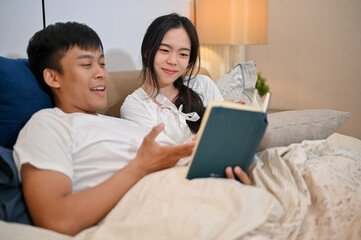 Obraz premium A happy young Asian couple in pajamas enjoys reading a book together before going to bed.