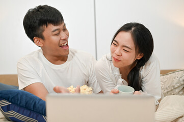 Happy Asian couple is having a fun time in bed, enjoying popcorn while watching a movie on a laptop.