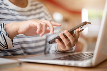A cropped image of a woman multitasking, using her smartphone while working on her laptop.