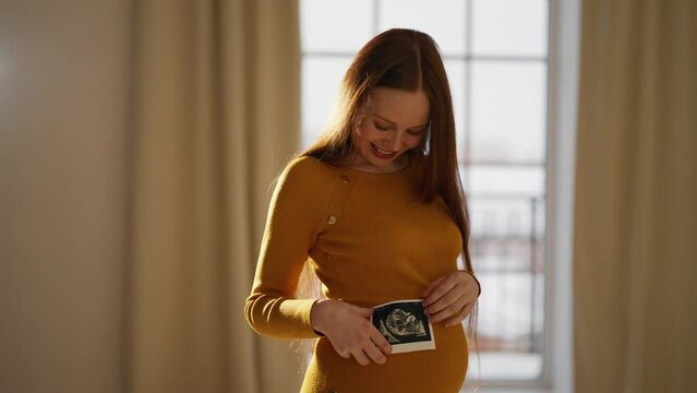Pregnant woman looking ultrasound report. Happy pregnant female watching her ultrasound report and touching her abdomen, admiring sonography picture of her unborn baby indoors. Expecting baby concept.
