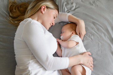 Tired mom lies on the bed next to her child, hugging him, he sucks a pacifier, falls asleep,...