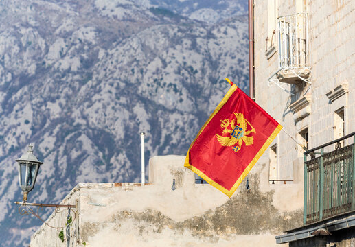 Montenegro's red flag, adorned with double-headed eagle, flutters against backdrop of mountainous landscape, beside vintage lantern and balcony. Kotor Bay, Perast, Montenegro