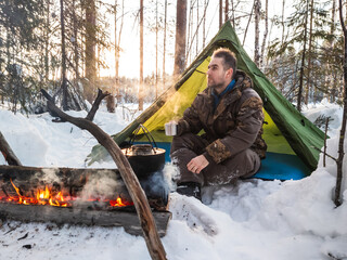 Man Enjoying a Warm Campfire Outside His Tent in a Snowy Forest at Dusk. Dressed for winter weather, he rests beside his tent pitched amidst a snowy landscape