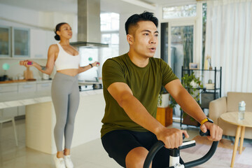 Fit man riding exercise bike when his girlfriend jumping with skipping rope