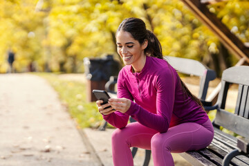 A fit sportswoman is sitting on a bench at city park and using her phone.