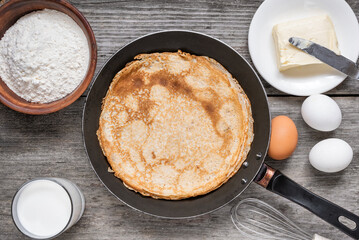 Frying pan with delicious crepes and ingredients for cooking on rustic wooden table. Thin pancakes in a pan with flour, eggs, milk and butter. Top view. Delicious breakfast