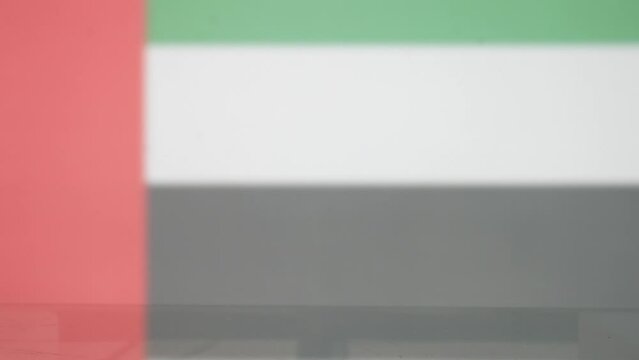 Close-up of a male hand taking a cup of tea with a tea bag tag of UAE flag from the glass table against the same blurred flag
