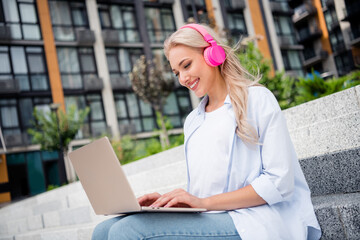 Photo of young pretty smiling satisfied woman in blue white shirt sitting stairs listen bluetooth...