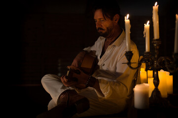 Side view of a man dressed in white enjoying playing Spanish guitar at home by candlelight, learning musical instrument, musician or singer singing new single song