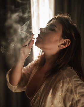 close-up of a beautiful girl smoking in her nightgown by the curtains, blowing smoke from her mouth.