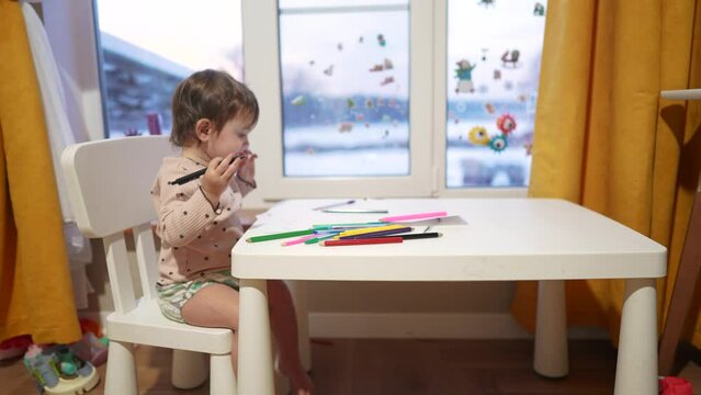 baby girl draws while sitting at a table by the window at home. happy family kid concept. baby daughter learns to dream draw with pencils on a sheet of paper indoors. development of fine motor skills