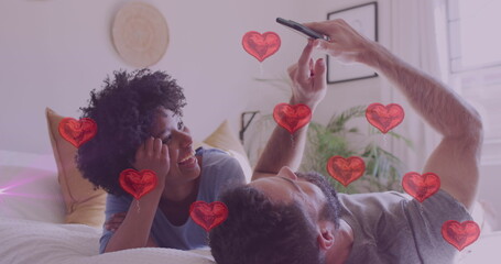 Image of hearts over diverse couple using smartphone in bed