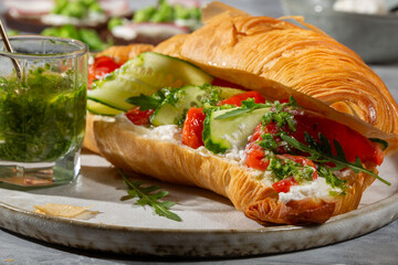 Croissant sandwich with cream cheese, smoked salmon, cucumber and arugula served with pesto on gray background.