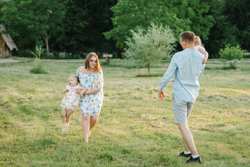 Naklejka na ściany i meble Concept of family spending time together. Mother, father hold hands children daughter. Running in green grass in nature on summer day. Mom, dad, and kids walking in park at sunset. Spring vacation.