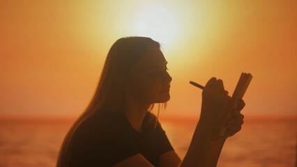 Silhouette of a young woman writing down her ideas in a notebook against the sunset background