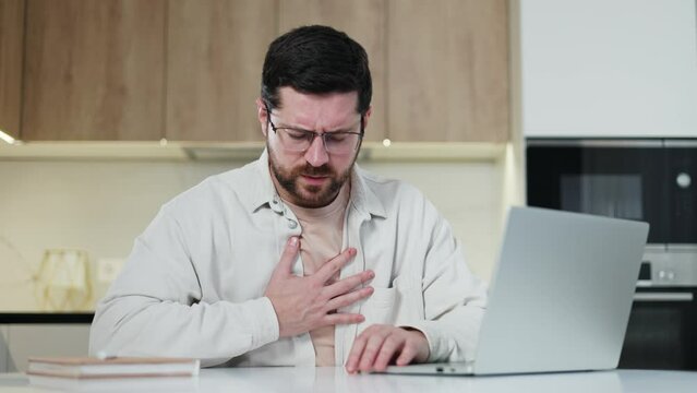 Caucasian freelancer with weak heart using wireless pc while sitting by desk on background of kitchen. Unhealthy guy experiencing cardiac stroke and holding sore spot why bending over table.