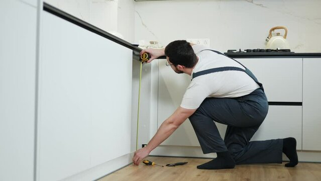 Workforce person measuring length and width of space under sink with building tape and writing indicators on clipboard. Busy man leaning on one knee and bending for checking inside size of cabinet.