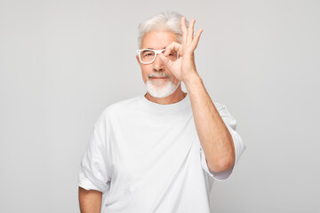 Senior man with glasses making OK gesture on a gray background.