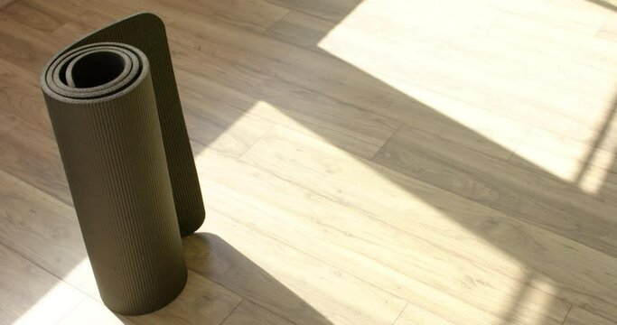 A rolled-up yoga mat stands on a wooden floor with sunlight casting shadows, with copy space