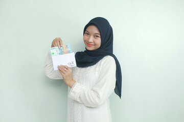 A young Asian Muslim woman holding an envelope and fifty thousand rupiah in cash with an expression...