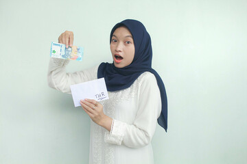 A young Asian Muslim woman holding an envelope and fifty thousand rupiah in cash with an expression...