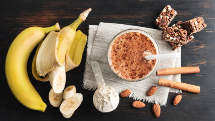 Glass with protein drink, healthy milkshake smoothie on wooden board with bananas, protein powder in measuring spoon, protein bar, almond nuts and cinnamon sticks, top view