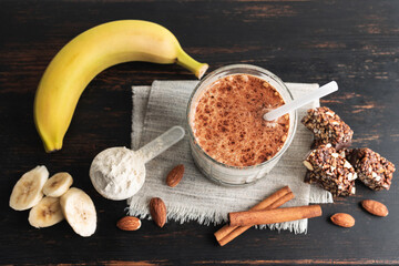Glass with protein drink, healthy milkshake smoothie on wooden board with bananas, protein powder in measuring spoon, protein bar, almond nuts and cinnamon sticks