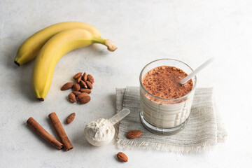 Fototapeta premium Glass with protein drink, healthy milkshake smoothie on white table with bananas, protein powder in measuring spoon, almond nuts and cinnamon sticks