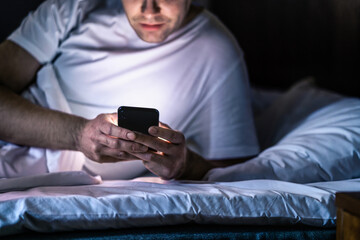 Man in bed with phone at night. Texting with smartphone before sleeping. Guy holding cellphone in hand. Dark home bedroom. Infidelity, cheating or working late online concept. Screen light on face. - obrazy, fototapety, plakaty