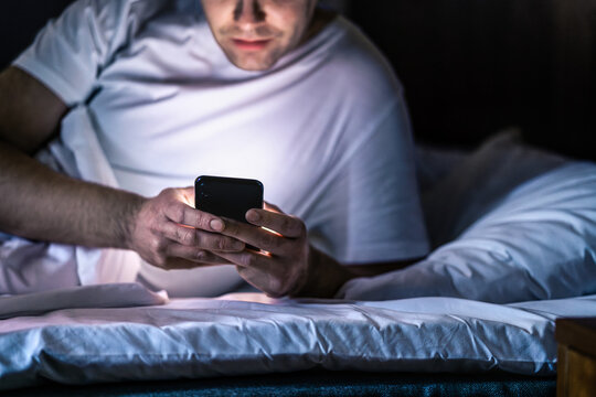 Man in bed with phone at night. Texting with smartphone before sleeping. Guy holding cellphone in hand. Dark home bedroom. Infidelity, cheating or working late online concept. Screen light on face.