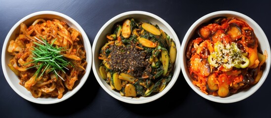 Three white bowls are filled with a flavorful medley of stir-fried kimchi and other classic Korean side dishes. The dishes showcase a variety of textures and colors, creating a visually appealing