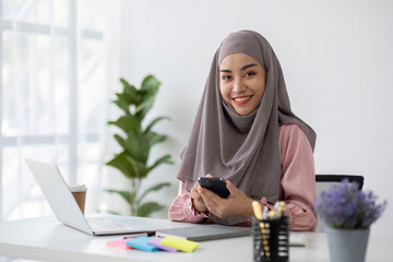 Smiling Muslim business woman Wearing a hijab while chatting online on a work desk in the office.